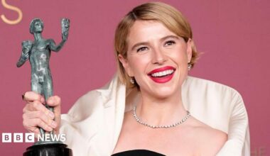 Buckley has short blonde hair and pink lipstick on. She is wearing a black and white dress and is holding up her grey award.