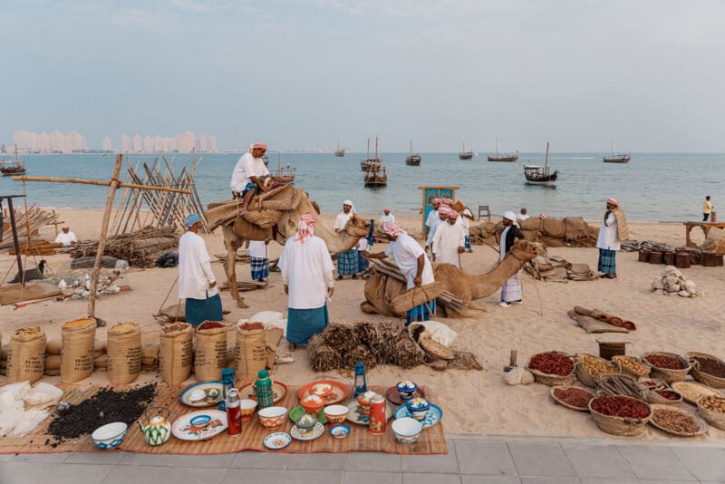 A seaside market scene with men in traditional Middle Eastern attire, camels, sacks of goods, spices, and colorful pottery displayed on the sand. Boats float on the water in the background under a cloudy sky.
