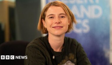 Jessie Buckley, who has short blonde hair and pierced ears with different earrings in. She is wearing a dark green knitted jumper and a bead necklace. She is smiling at the camera and sitting in front of a Desert Island Discs backdrop