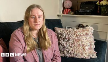 A woman in a pink shirt with long blonde hair sits on a dark green sofa. There are cushions and a blanket next to her. Behind her you can see a typewriter and some roses on a shelf.