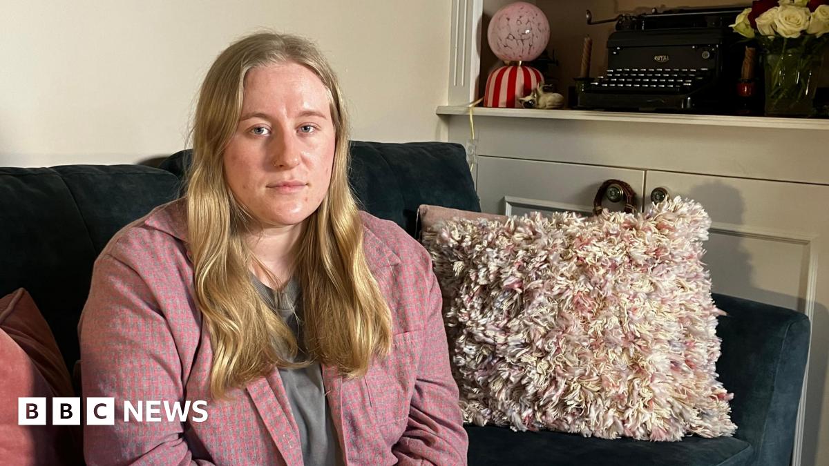 A woman in a pink shirt with long blonde hair sits on a dark green sofa. There are cushions and a blanket next to her. Behind her you can see a typewriter and some roses on a shelf.