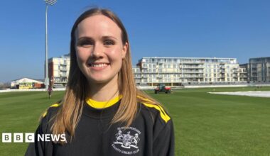 A women is smiling at the camera. She is wearing a Gloucestershire County Cricket Club branded t-shirt which is black with yellow stripes across the shoulders and around the neck. She has mid-length, light brown hair. She is stood on a cricket ground.