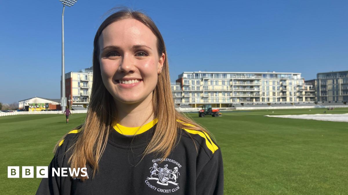 A women is smiling at the camera. She is wearing a Gloucestershire County Cricket Club branded t-shirt which is black with yellow stripes across the shoulders and around the neck. She has mid-length, light brown hair. She is stood on a cricket ground.