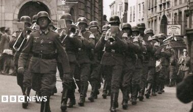 A German solider holding a rifle and wearing a tin helmet is standing on a cliff and looking out to sea
