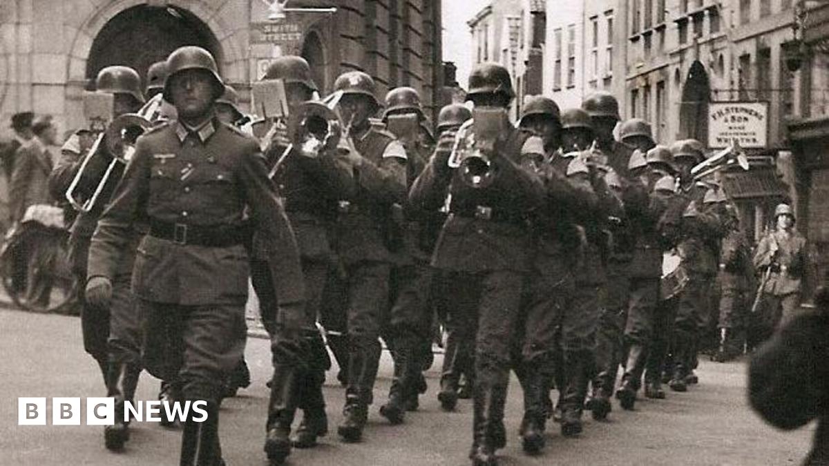 A German solider holding a rifle and wearing a tin helmet is standing on a cliff and looking out to sea