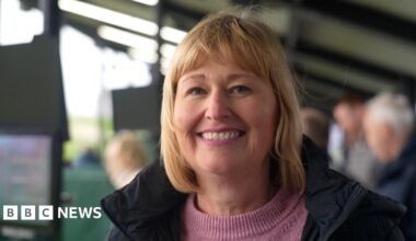 Andy is wearing a black and grey zip-up sports top. He is unshaven and is standing outside at a golfing centre and/or driving range.
