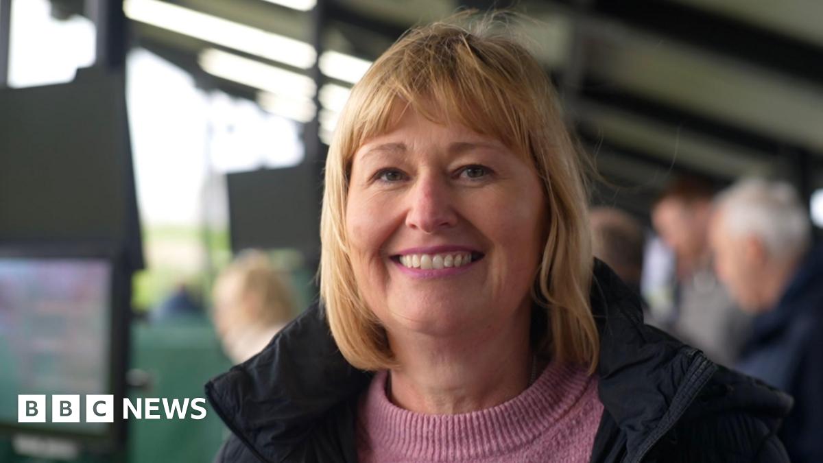 Andy is wearing a black and grey zip-up sports top. He is unshaven and is standing outside at a golfing centre and/or driving range.