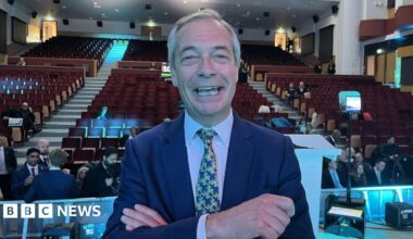 Nigel Farage smiling with an open mouth looking directly at the camera, wearing suit and tie and his arms crossed. He is standing in front of hundreds of auditorium seats, some of which are filled with people wearing suits and formal wear.