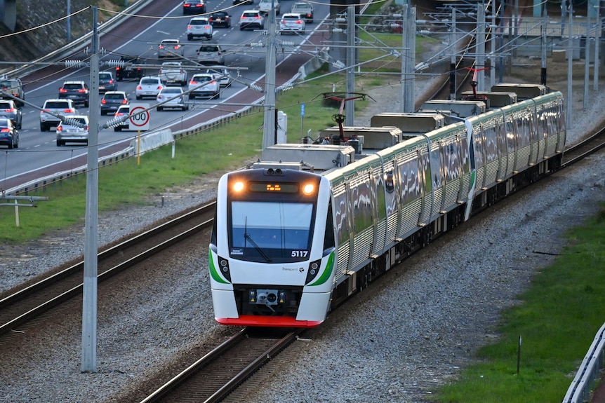 A Transperth passenger train in daylight moving on a railway track near a freeway.