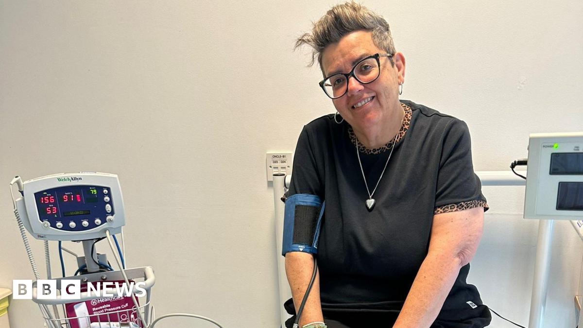 An older woman with short grey hair sits in a hospital room, attached to a heart rate monitor machine. She wears glasses, silver hooped earrings, a black t-shirt with a leopard-print trim around the sleeves and neckline.