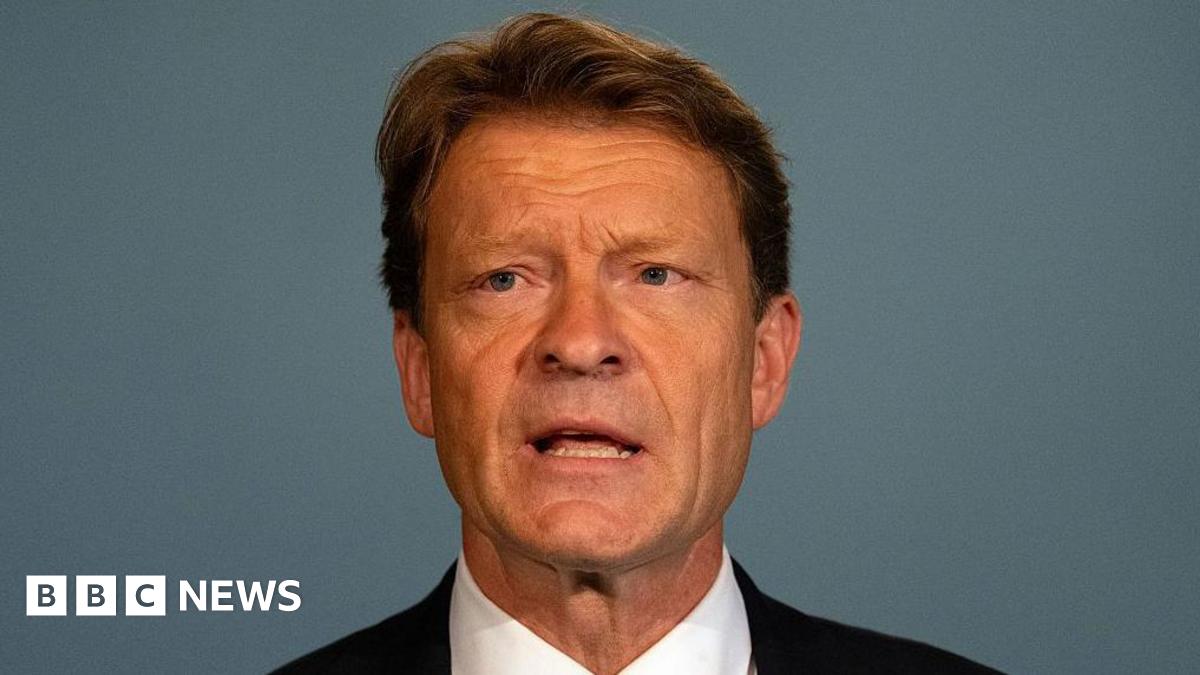 Richard Tice giving a speech. Tice has short brown hair and wears a black suit jacket, white shirt, and a black jacket. mall microphones positioned in front of them on the table.