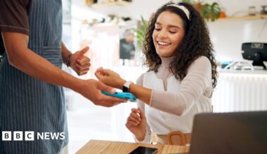 A curly brown haired woman uses her watch for card payment in a cafe - she has a laptop in front of her. her phone lies on the table. She wears a white top, brown belt and white headband.