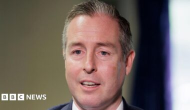 A man with short cropped dark and grey hair is seen head on, the tops of his shoulders are visible in the image. He has his mouth slightly open, as if he is in the middle of speaking. He is wearing a dark jacket and a white shirt. The background is blurred but it appears to be an indoor setting.