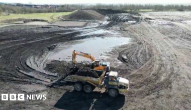A lorry and a yellow digger at work on the muddy site of the new hospital. There are large puddles.