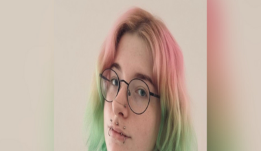 Portrait photo of young woman with pink and green hair, round glasses and a number of lip piercings.