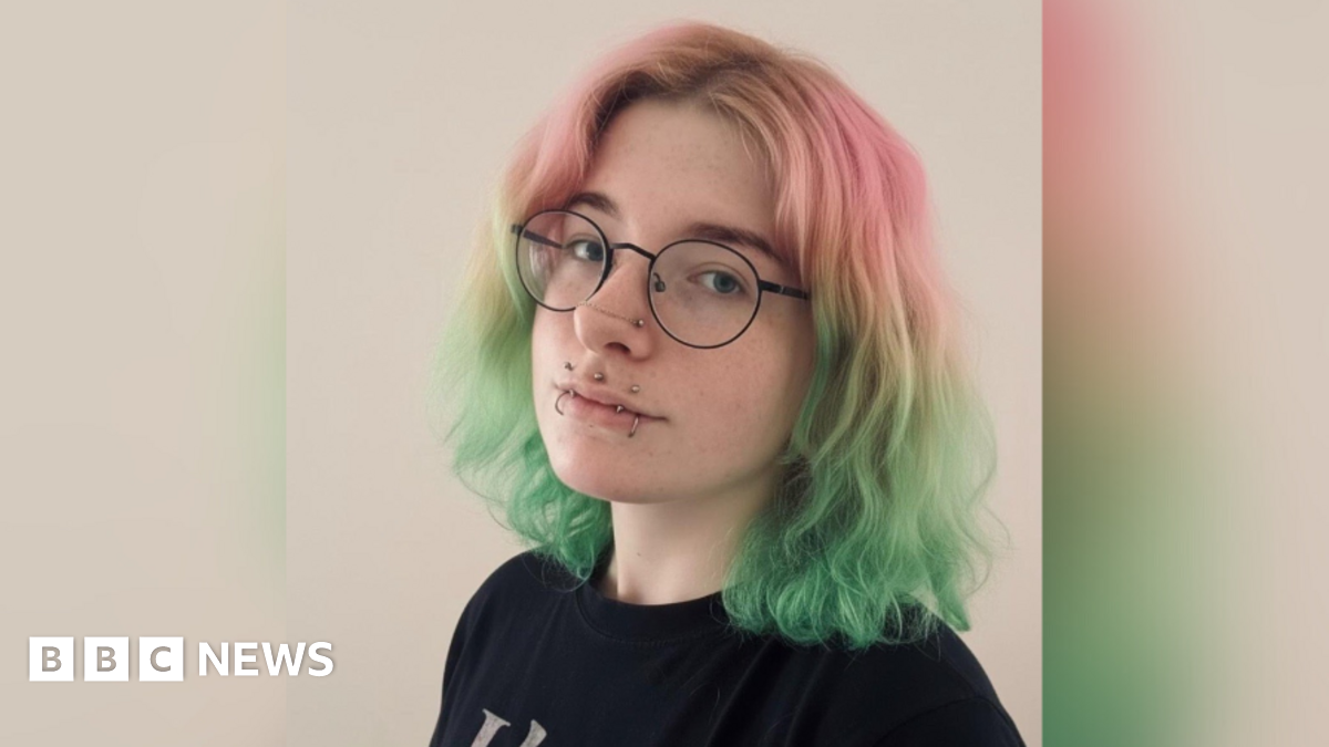 Portrait photo of young woman with pink and green hair, round glasses and a number of lip piercings.