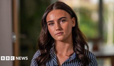 Sasha looking at the camera with a serious expression. She has long brown curly hair and wears a navy and white shirt. She is sat in her living room with the background blurred behind her.