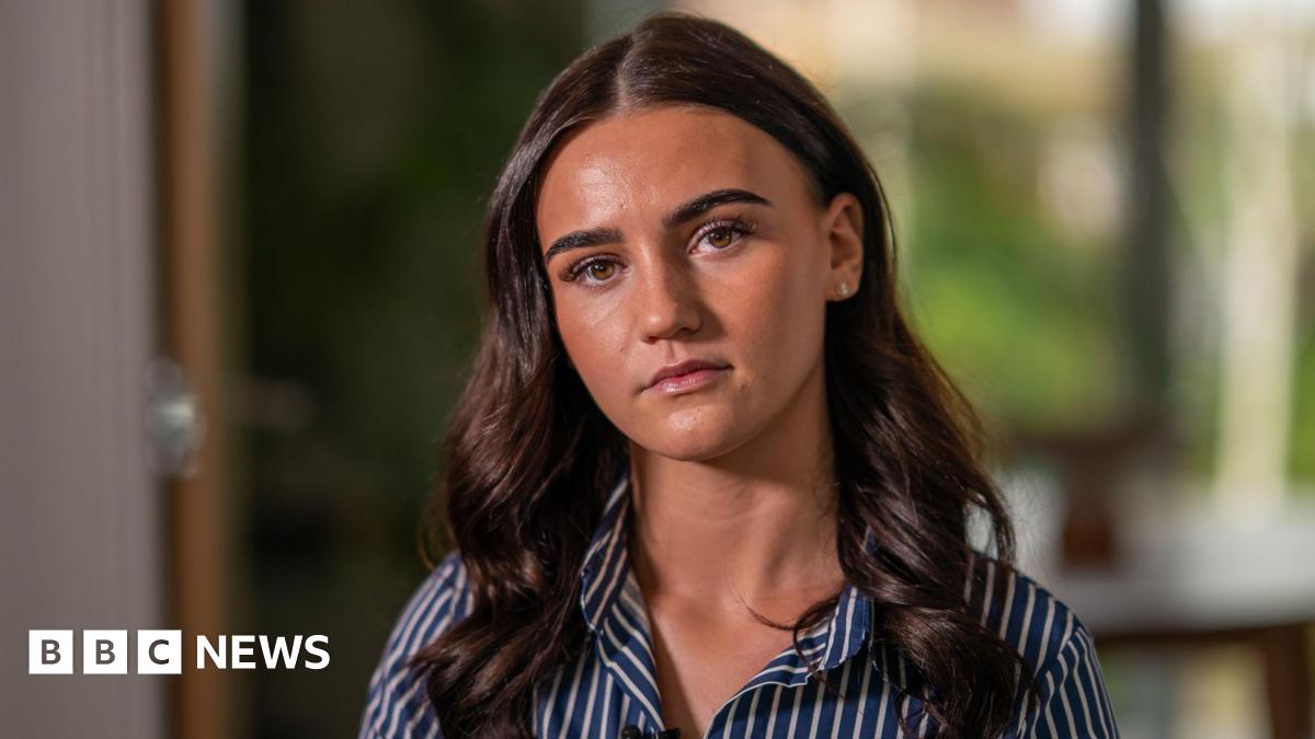 Sasha looking at the camera with a serious expression. She has long brown curly hair and wears a navy and white shirt. She is sat in her living room with the background blurred behind her.