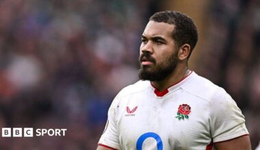 Close-up of England's Ollie Lawrence. He has short black hair and a beard, and is wearing a white shirt which has the English team's crest - a rose - on the left breast and manafacturer's logo on the right.