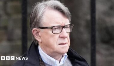 Lord Mandelson wearing glasses, a dark coloured coat, a grey jumper and a shirt.