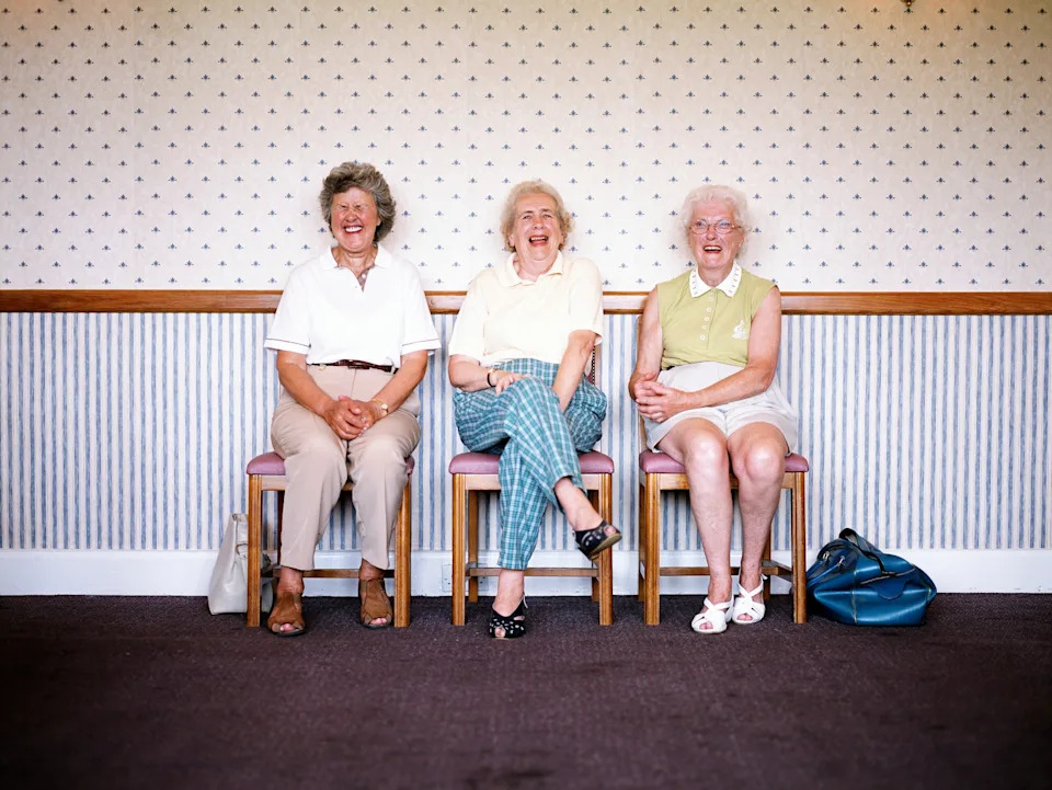 Three elderly women sit on a bench in a room with patterned wallpaper, laughing and enjoying each other's company