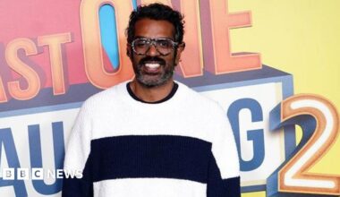 Romesh Ranganathan is seen during the photocall for Prime Video's Last One Laughing. He is wearing a black and white jumper and smiling and the logo for the programme is behind him.
