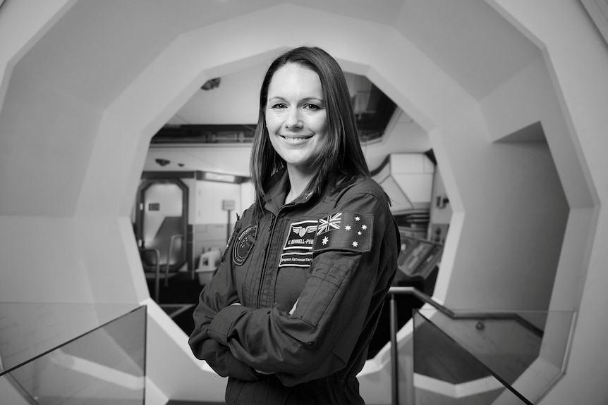 A smiling woman in a space suit standing in front of a octagonal doorway.  