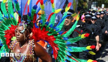 A brightly-feathered parade participant stands in front of a police line during the Notting Hill Carnival on August 25, 2025