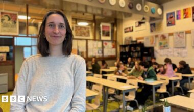 A brunette woman with a bob smiles at the camera in front of a classroom of teenagers. She is smiling and wearing a grey jumper. There are about a dozen children in the class