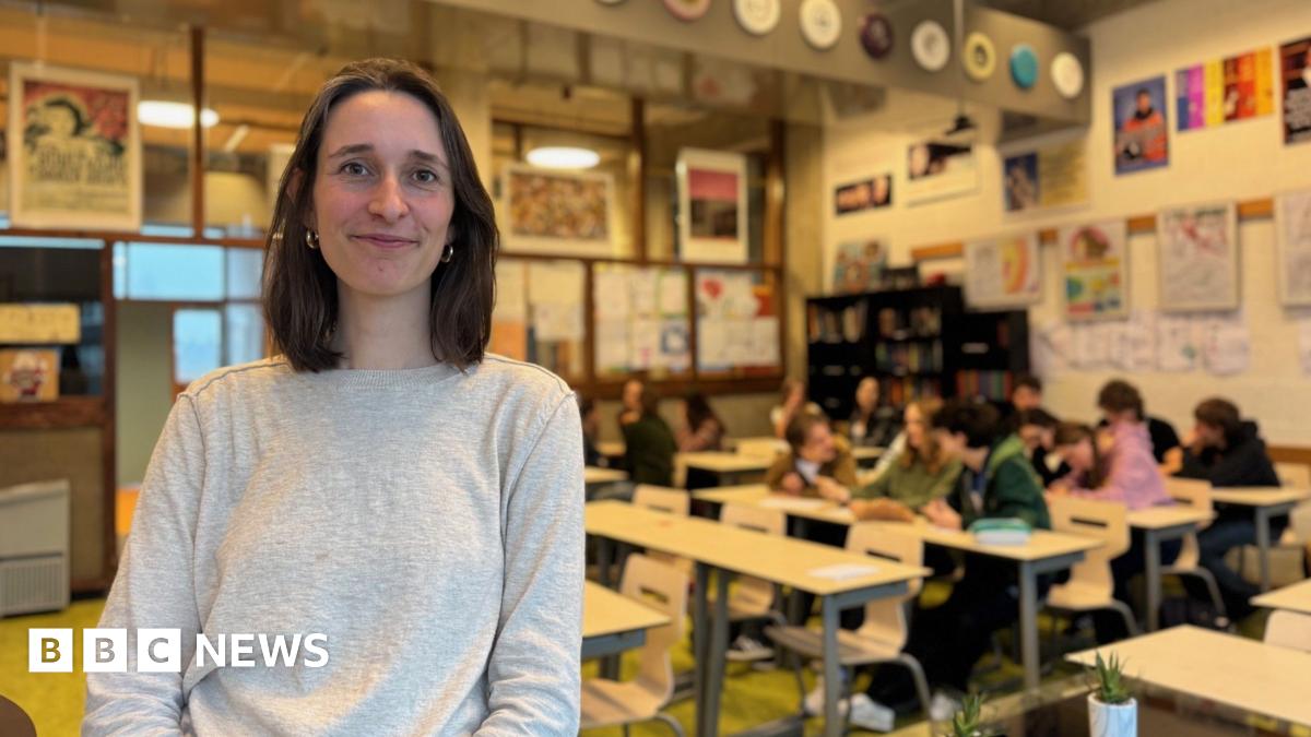 A brunette woman with a bob smiles at the camera in front of a classroom of teenagers. She is smiling and wearing a grey jumper. There are about a dozen children in the class
