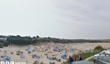 The image shows a wide sandy beach filled with groups of people sitting close to colourful windbreaks spread across the sand. Some people are walking along the shoreline while others are sitting or lying down on towels and beach chairs. There are a few small tents and beach shelters scattered around.
