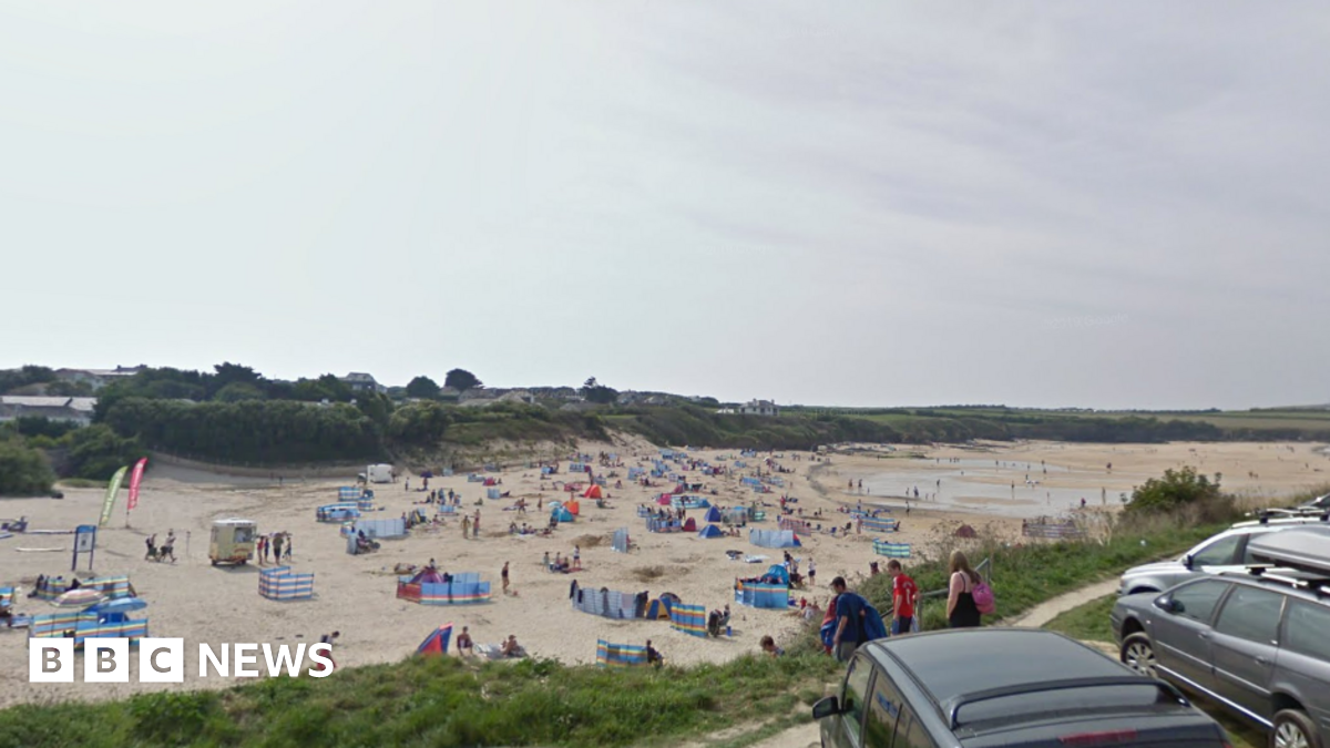 The image shows a wide sandy beach filled with groups of people sitting close to colourful windbreaks spread across the sand. Some people are walking along the shoreline while others are sitting or lying down on towels and beach chairs. There are a few small tents and beach shelters scattered around.
