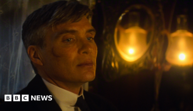 Cillian Murphy, playing Tommy Shelby in a film still, looking away from the camera. He has a scar on his face and lights glowing yellow behind him. His hair is slightly grey.