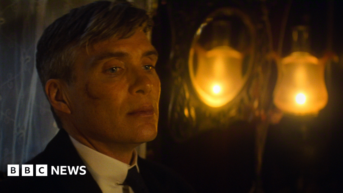Cillian Murphy, playing Tommy Shelby in a film still, looking away from the camera. He has a scar on his face and lights glowing yellow behind him. His hair is slightly grey.