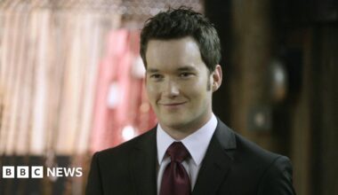A wall covered in pictures and a string of Welsh flags. At the top a board reads Ianto's Shrine in white writing. The pictures are attached to a rusty metal grid.