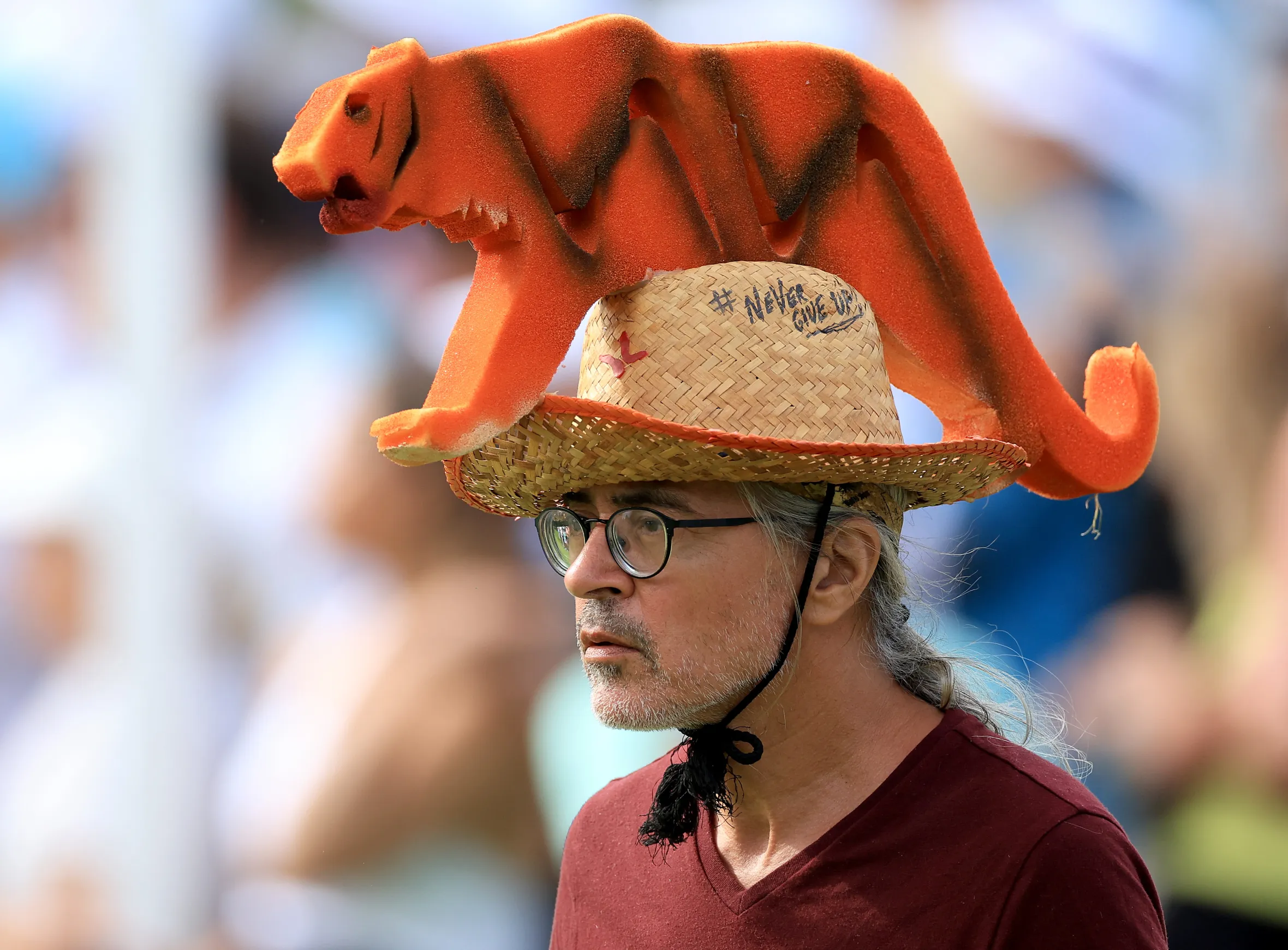 A fan at the PGA Championship wearing a straw hat with an orange foam tiger on top and "Never Give Up" written on it.
