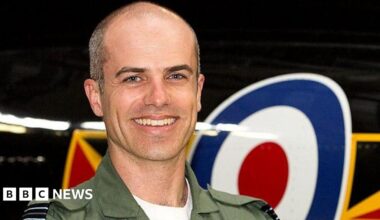 Close up image of Geoff Corser smiling at the camera standing in front of an RAF plane. He is wearing khaki green uniform with a white t-shirt. Geoff has very short grey hair.