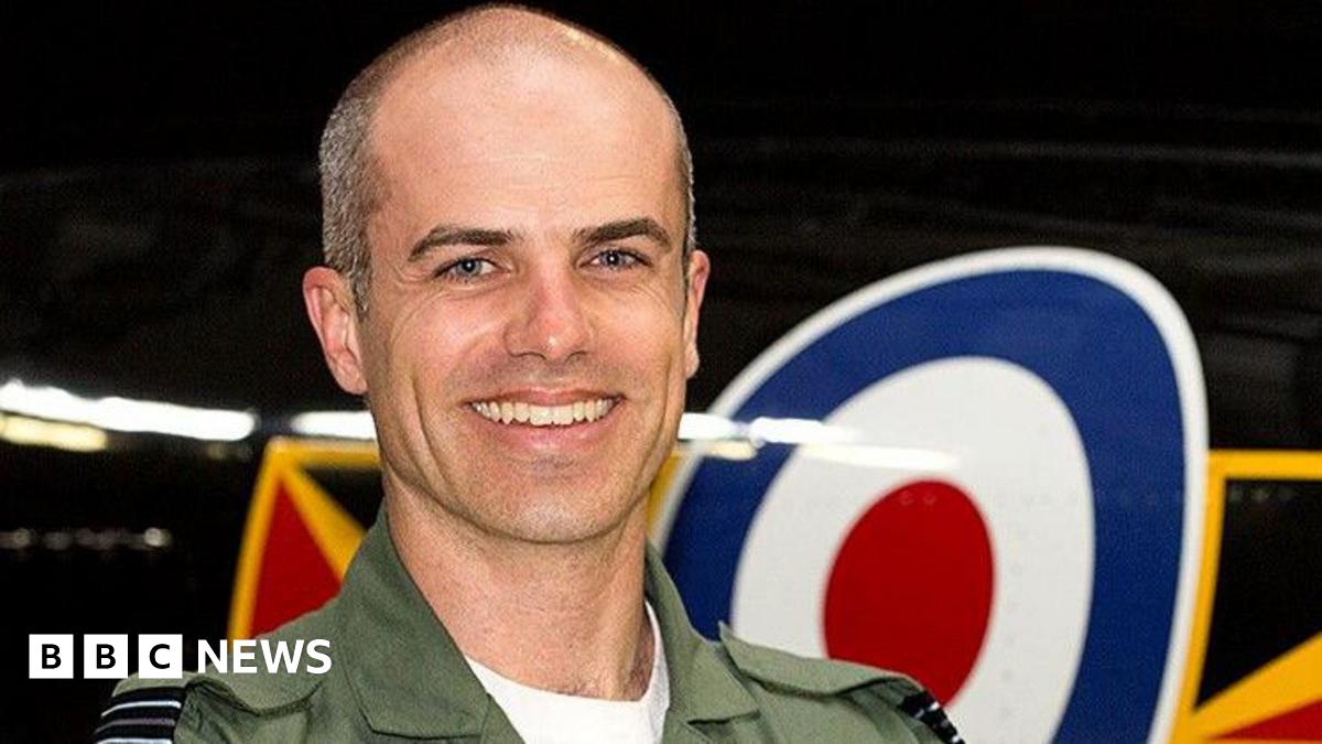 Close up image of Geoff Corser smiling at the camera standing in front of an RAF plane. He is wearing khaki green uniform with a white t-shirt. Geoff has very short grey hair.