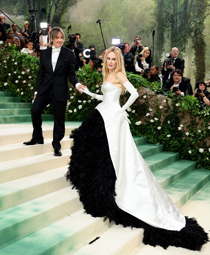 Nicole Kidman in black and white gown on staircase at event, surrounded by photographers and greenery background. Nicole Kidman in black and white gown on staircase at event, surrounded by photographers and greenery background.