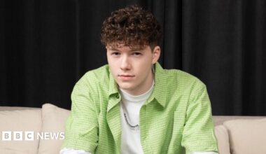 Sam Meyer, who is wearing a white T-shirt with a lime green shirt and a silver chain, is staring directly at the camera while sitting on a cream couch.