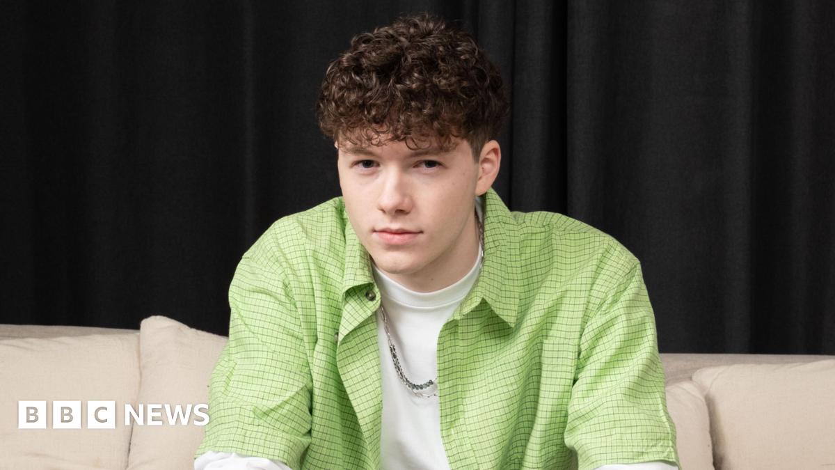 Sam Meyer, who is wearing a white T-shirt with a lime green shirt and a silver chain, is staring directly at the camera while sitting on a cream couch.