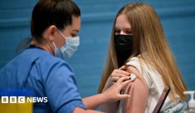 A vaccination setting taking place indoors. A healthcare worker wearing a blue medical uniform is administering an injection into the upper arm of another individual.