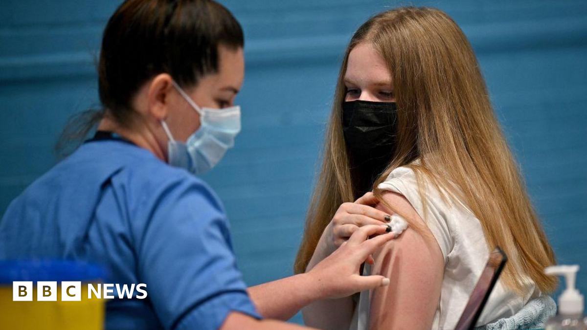 A vaccination setting taking place indoors. A healthcare worker wearing a blue medical uniform is administering an injection into the upper arm of another individual.