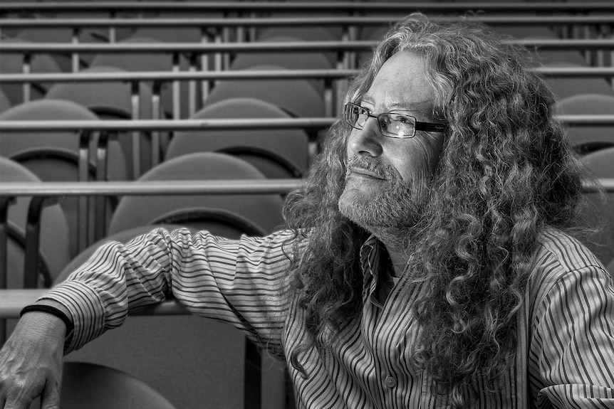 A black-and-white photo of a man with long curly hair and spectacles, sitting in an empty lecture theatre. 