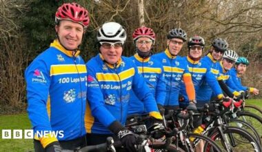 The cyclists are lined up with their bikes and wear bright blue and yellow cycling tops with 100 laps for Rob on them.  They wear helmets of varying colours.