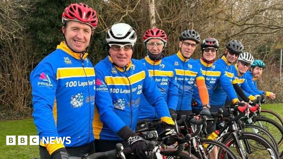 The cyclists are lined up with their bikes and wear bright blue and yellow cycling tops with 100 laps for Rob on them.  They wear helmets of varying colours.