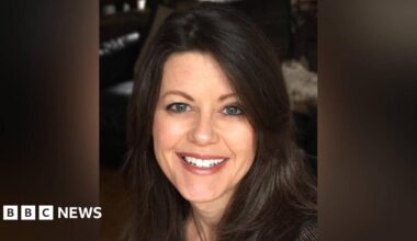 A head shot of Amanda who is looking at the camera and smiling, showing her top front teeth.  She has blue eyes and dark brown shoulder lenghth brown hair. The background is blurred and dark