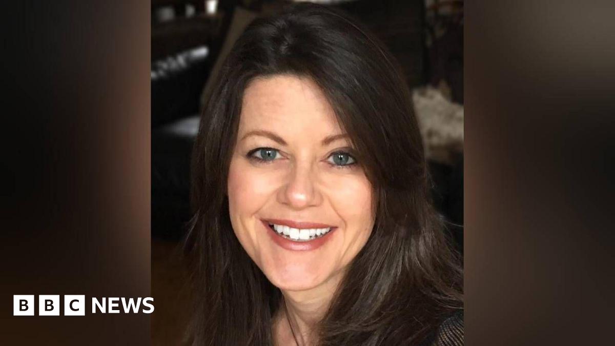 A head shot of Amanda who is looking at the camera and smiling, showing her top front teeth.  She has blue eyes and dark brown shoulder lenghth brown hair. The background is blurred and dark