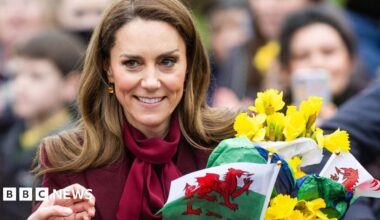 Catherine wearing burgundy carrying yellow daffodils and Welsh flags as people take pictures in the background