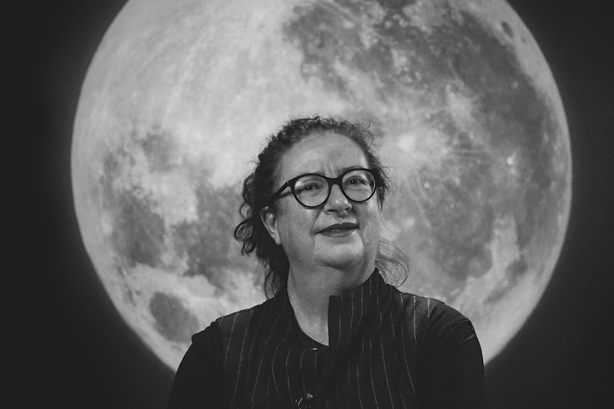 A smiling woman wearing spectacles stands with her arms folded, in front of a full moon.